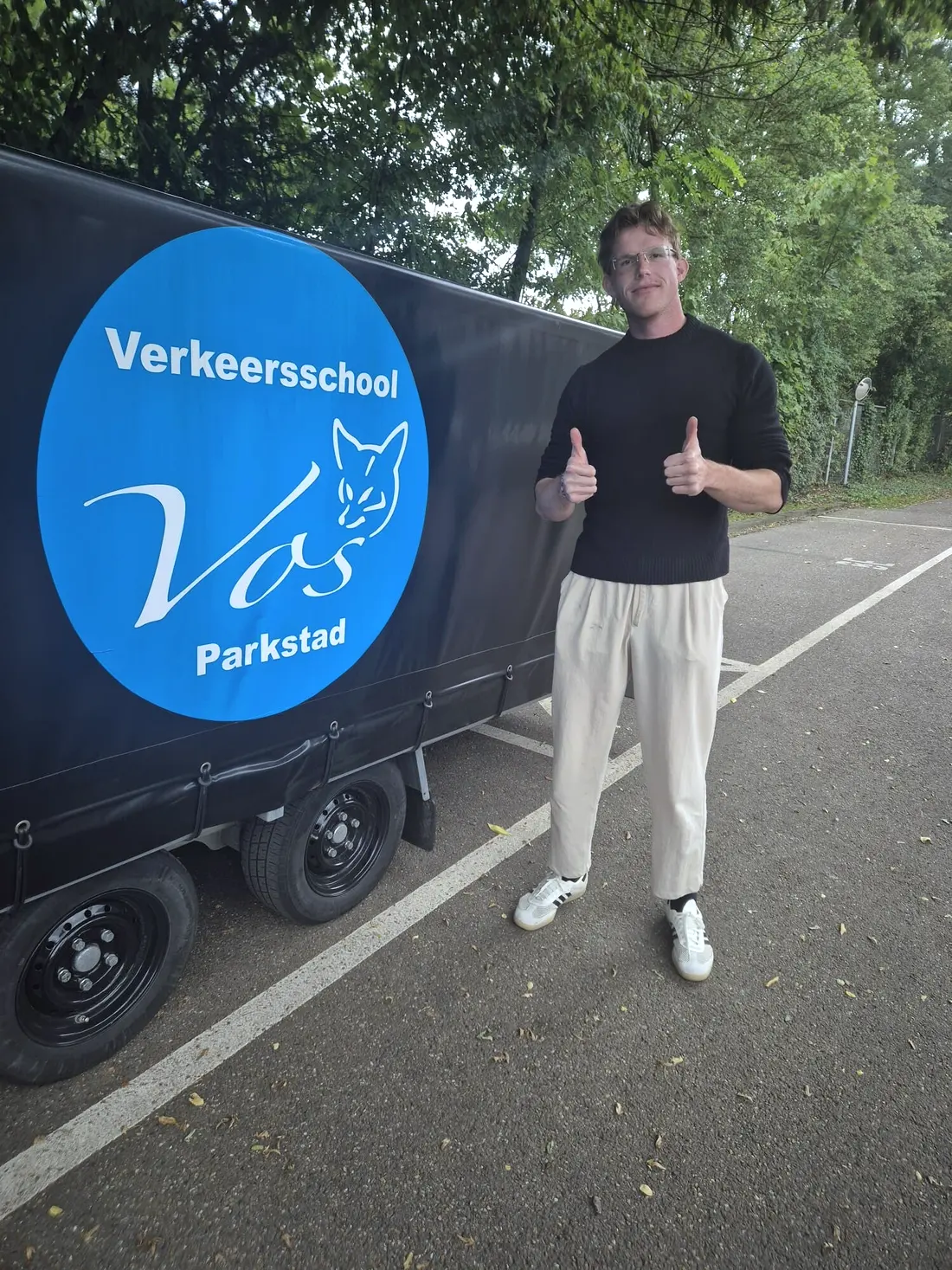 Geslaagde leerling met de duim omhoog bij de aanhanger van Verkeersschool Vos