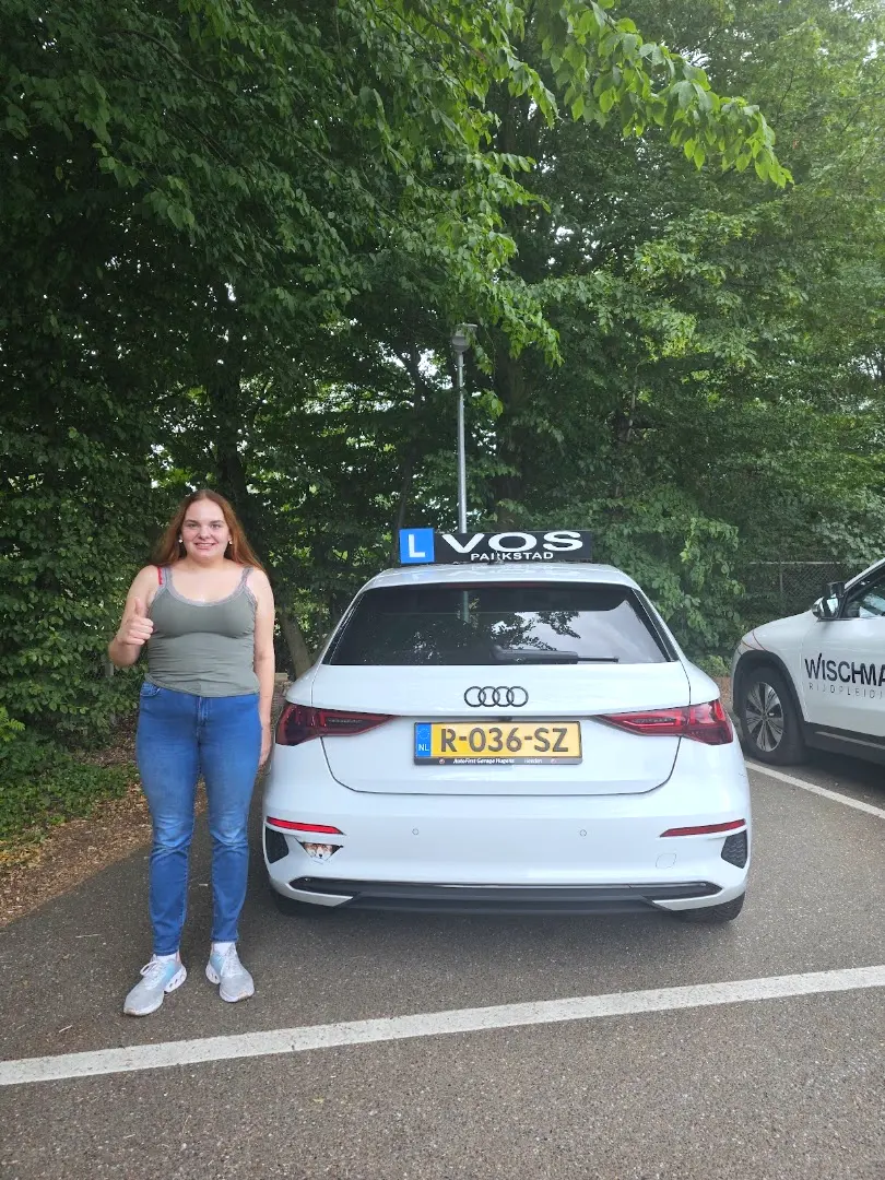 Geslaagde leerling steekt haar duim op naast de witte lesauto van Verkeersschool Vos, op dezelfde parkeerplaats